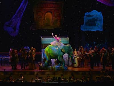 Opera Siam stage production features a performer atop a colorful elephant set piece with orchestra and ensemble.