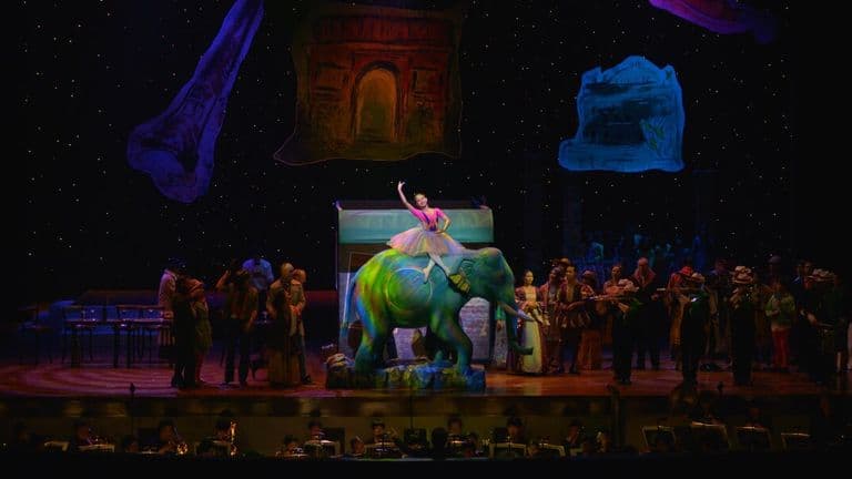 Opera Siam stage production features a performer atop a colorful elephant set piece with orchestra and ensemble.