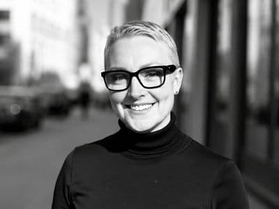 Jeannette Papadakis smiling outdoors in a black turtleneck and glasses.