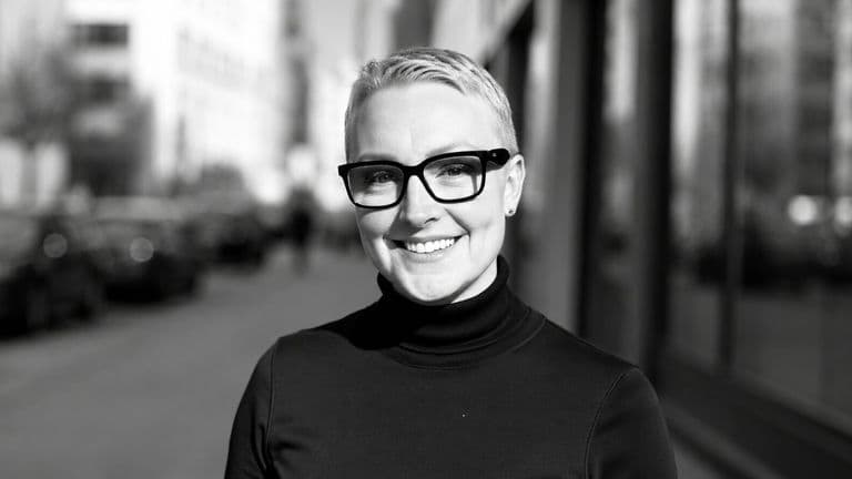 Jeannette Papadakis smiling outdoors in a black turtleneck and glasses.
