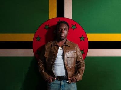 Jordn in a brown leather jacket standing in front of the Dominica flag.