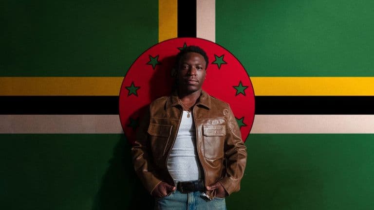 Jordn in a brown leather jacket standing in front of the Dominica flag.