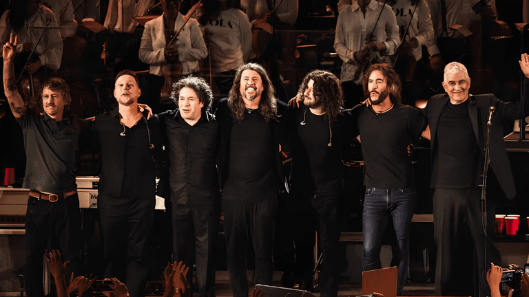 Seven musicians in black clothing stand on stage, waving to an audience.