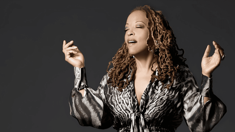 A woman with curly hair sings passionately, wearing a stylish animal-print blouse