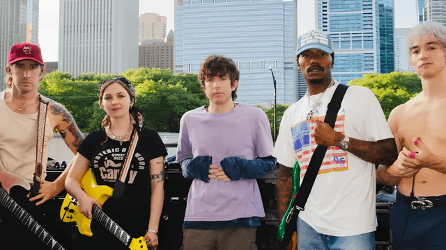 Five musicians pose outside with guitars in front of a cityscape background