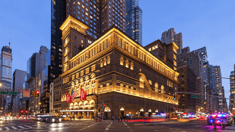 Carnegie Hall illuminated at dusk, surrounded by skyscrapers and city traffic