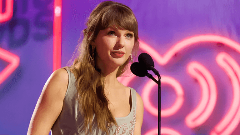 A woman with long hair speaks at a microphone on stage with neon lights behind her