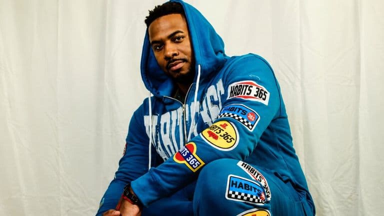 Portrait of rapper Ju-Lion The Voice wearing a blue tracksuit with racing patches against a white backdrop.