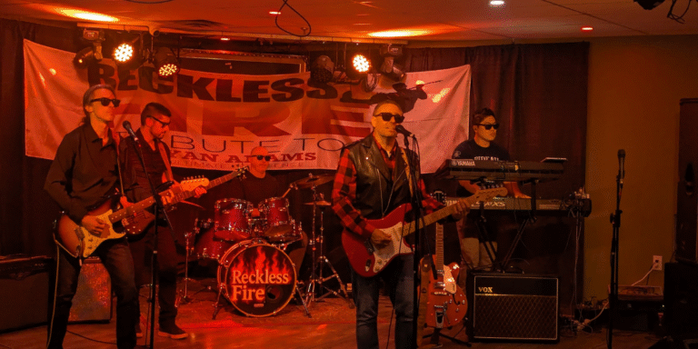 Reckless Fire: Ottawa’s Bryan Adams Tribute Band Capturing the Heart and Spirit of a Rock Legend "Band performing live with instruments, stage lighting, and a banner featuring 'Reckless Fire' at a tribute concert."