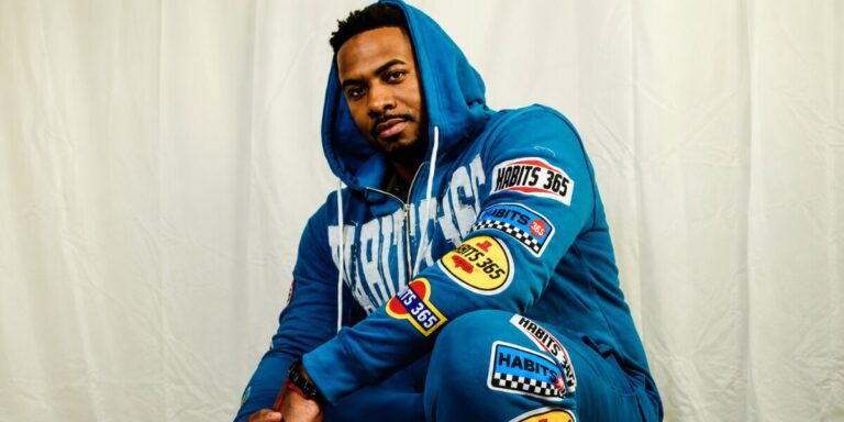 Ju-Lion: A Brand And A Voice Portrait of rapper Ju-Lion The Voice wearing a blue tracksuit with racing patches against a white backdrop.