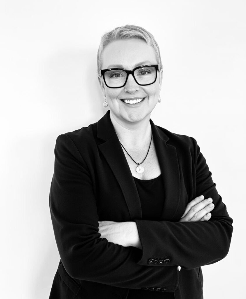Jeannette Papadakis smiling with folded arms in a black blazer and glasses.