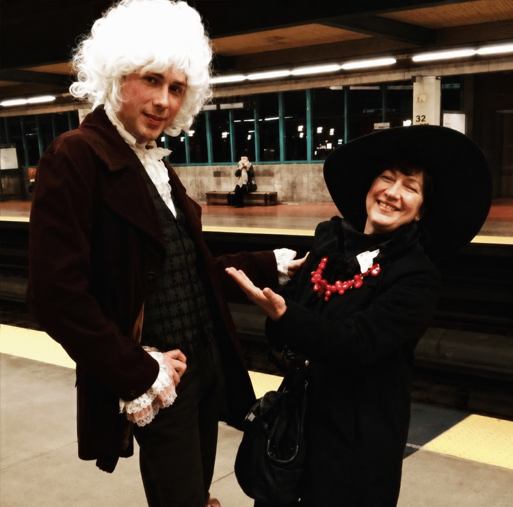 Two people in 18th-century-inspired attire posing on a subway platform.