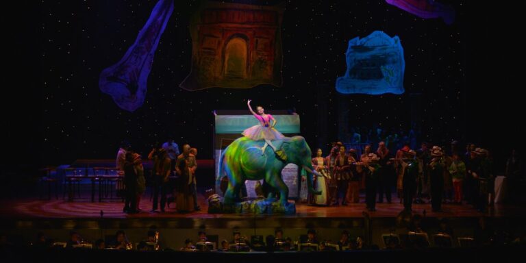 Opera Siam stage production features a performer atop a colorful elephant set piece with orchestra and ensemble.