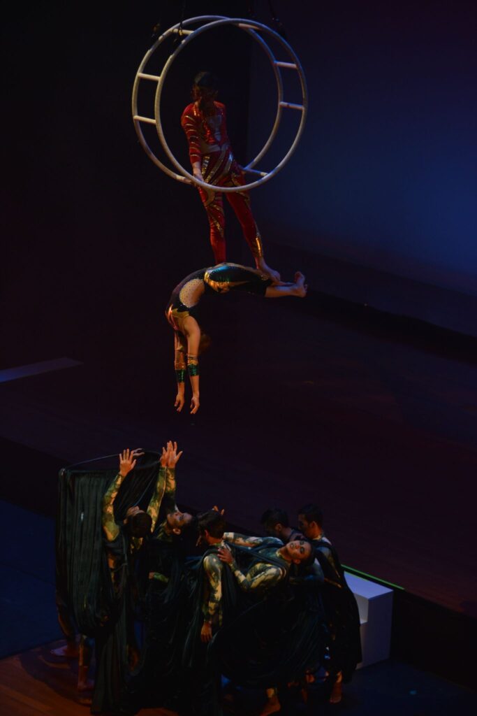 Acrobatic performers balance beneath a suspended circular apparatus during a dramatically lit Opera Siam stage production.