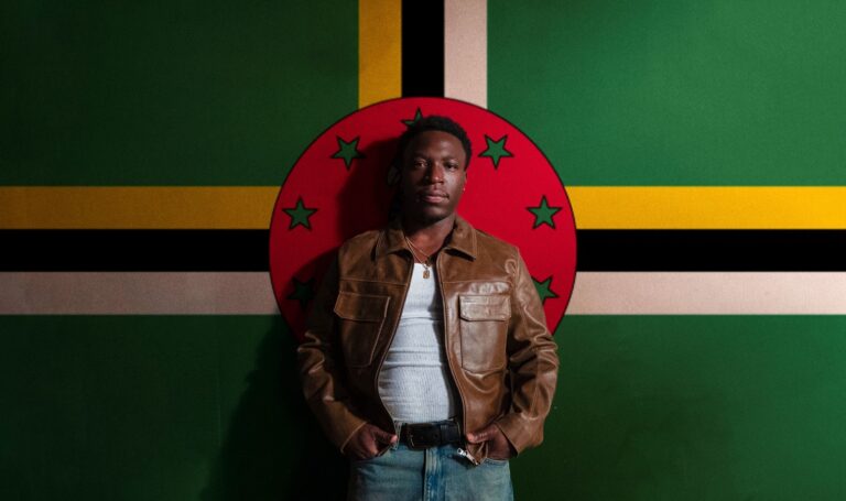 From Dominica to LA: The Rise of Jordn Jordn in a brown leather jacket standing in front of the Dominica flag.